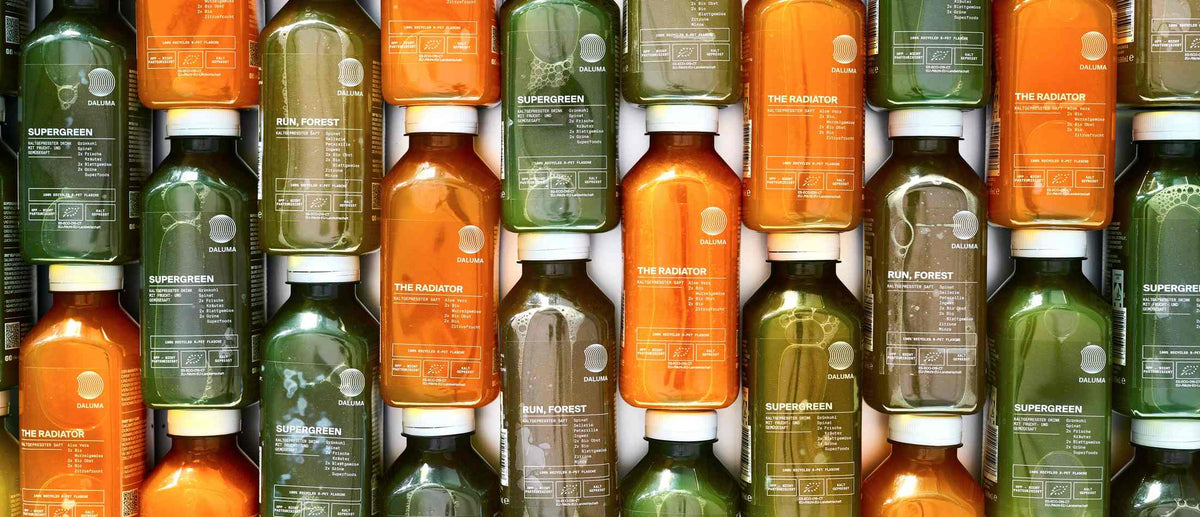 Viele DALUMA Saftflaschen in Grün und Orange dicht nebeneinander gestapelt, mit den Sorten SUPERGREEN, RUN, FOREST und THE RADIATOR