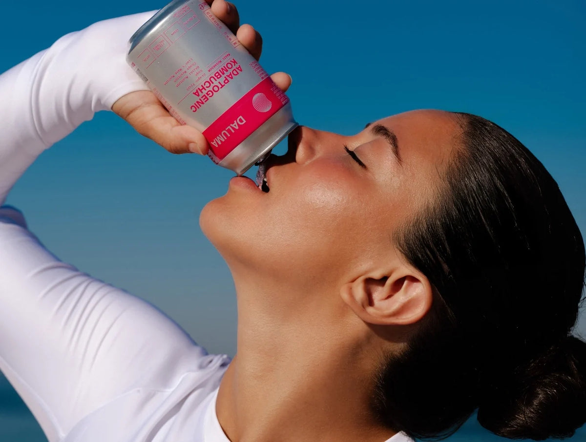 Frau im weißen Sportoutfit trinkt mit geschlossenen Augen aus einer Dose DALUMA Adaptogenic Kombucha vor strahlend blauem Himmel