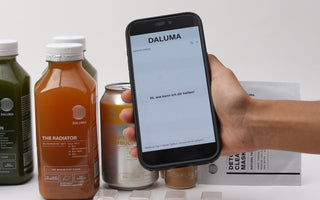 Smartphone mit dem DALUMA Health Coach Chat vor kaltgepressten Säften, Kombucha und Detox Clearing Mask.