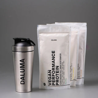Ein Edelstahl-Shaker mit der Aufschrift DALUMA steht neben drei weißen Beuteln mit DALUMA Vegan Performance Protein vor einem schlichten grauen Hintergrund.