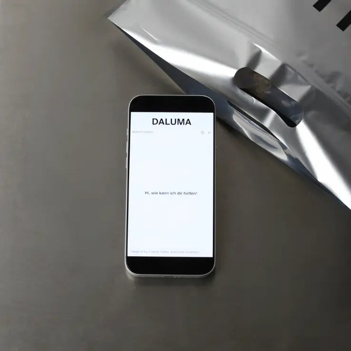 Ein Smartphone mit dem Schriftzug DALUMA und einer Grußbotschaft auf dem Display liegt auf einer grauen Fläche neben einer teilweise sichtbaren metallischen Verpackungstüte.