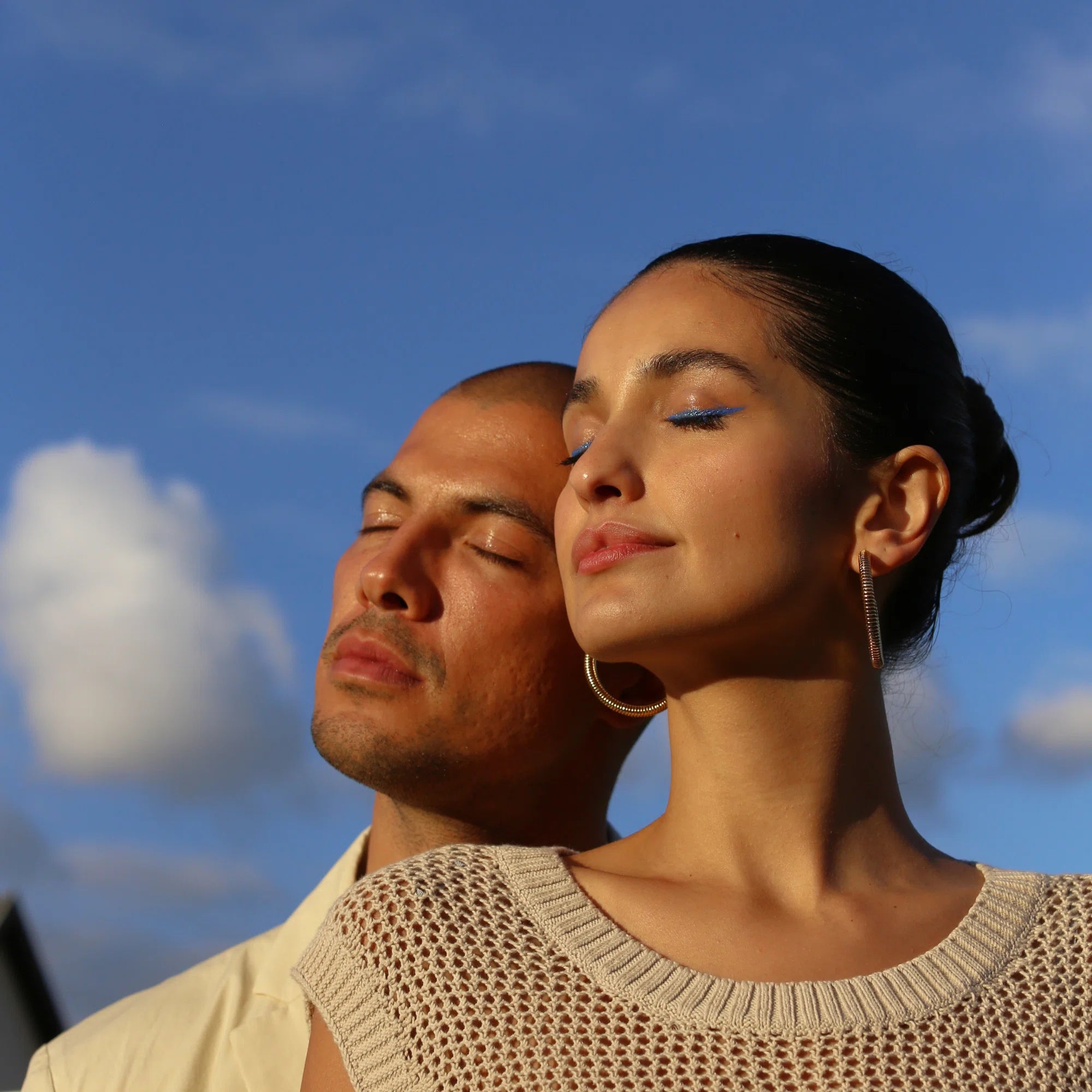 Ein Mann und eine Frau stehen friedlich im Freien, die Augen geschlossen im Sonnenlicht, als ob sie die Vorteile von DALUMA 360 DAILY ESSENTIALS SUPPLEMENT unter einem blauen Himmel mit Wolken genießen würden.