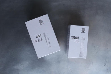 Zwei weiße rechteckige DALUMA-Ergänzungsdosen, darunter 360 DAILY ESSENTIALS SUPPLEMENT, ruhen auf einer Metalloberfläche. Beide haben ein minimalistisches Design, ein rundes Logo, Inhaltsstofflisten und heben natürliche Inhaltsstoffe hervor.