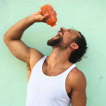 Ein Mann in einem weißen Tank-Top hält eine orangefarbene Getränkeflasche über den Mund und neigt den Kopf nach hinten, als würde er das Getränk einschenken, während er vor einem hellgrünen Hintergrund steht.
