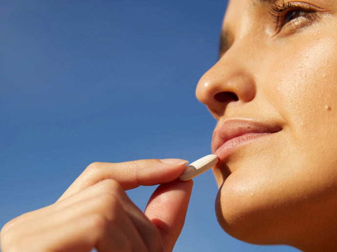 Nahaufnahme des Gesichts einer Person, die eine weiße Tablette oder Pille vor einem strahlend blauen Himmel an die Lippen hält, was darauf hindeutet, dass sie im Begriff ist, das Medikament oder das Ergänzungsmittel einzunehmen.