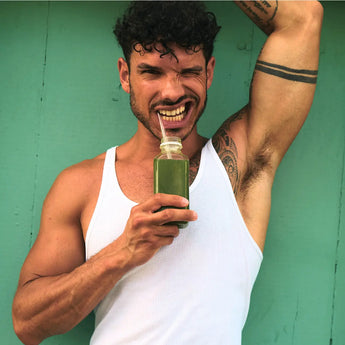 Ein Mann in einem weißen Tank-Top lächelt und zwinkert, während er eine Flasche grünen Saft mit einem Strohhalm vor einer blaugrünen Wand hält. Ein Arm ist angehoben und zeigt Tätowierungen auf seinem Arm und seiner Schulter.