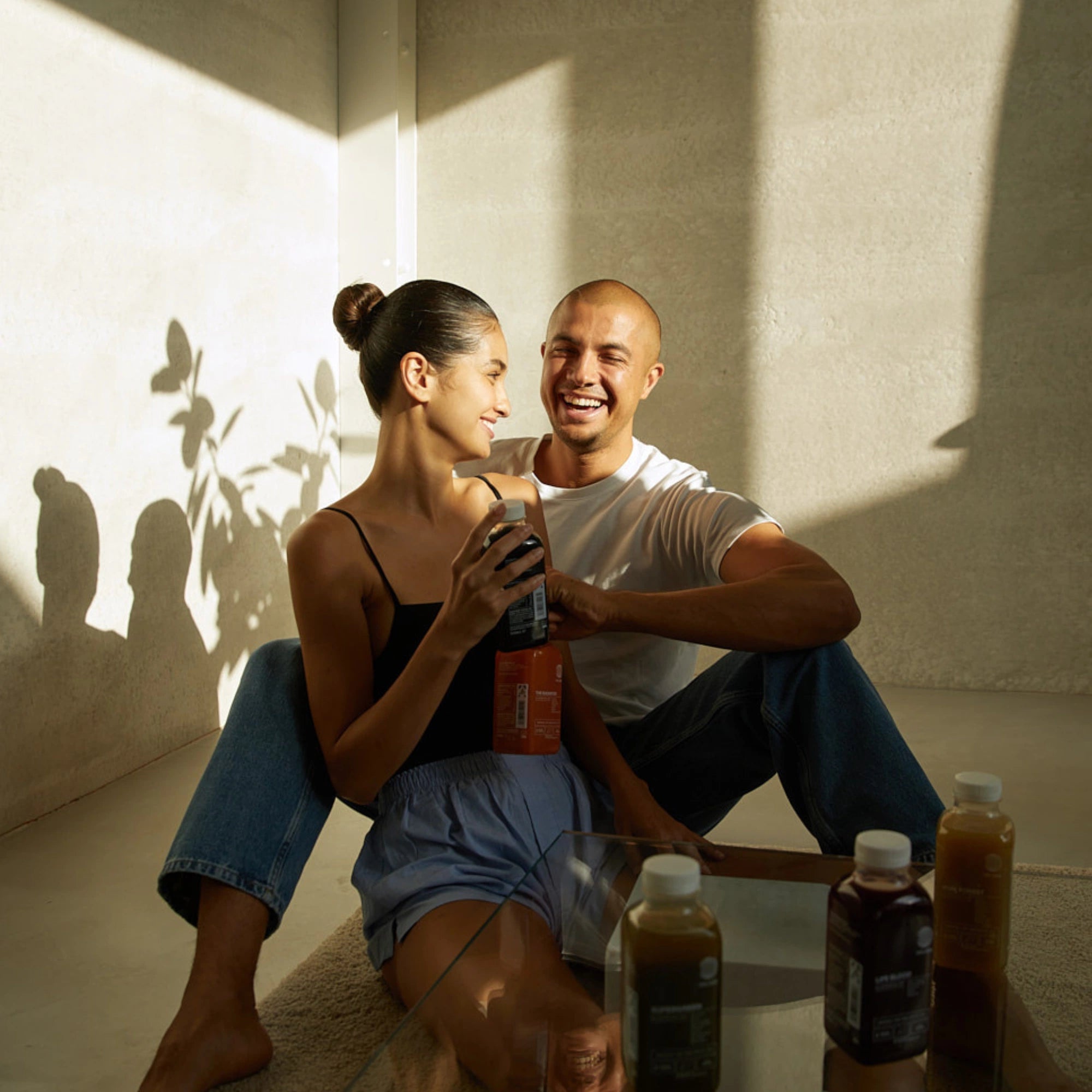 Ein lächelndes Paar entspannt sich gemeinsam auf dem Boden im Sonnenlicht. Die Frau hält eine Flasche DALUMA BESSERE SAFTKUR 7 TAGE in der Hand, auf dem Glastisch daneben stehen weitere Getränkeflaschen von DALUMA. Die minimalistische Dekoration umfasst Schatten und Pflanzen.