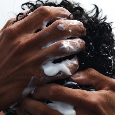 Nahaufnahme von Händen, die das HAIR CARE DUO Shampoo von Daluma-shop in dunkles, lockiges Haar einmassieren - weiße Seifenlauge und Finger sind zu sehen - ein veganer Haarpflege-Moment für kräftiges Haar vor einem unscharfen, hellen Hintergrund.