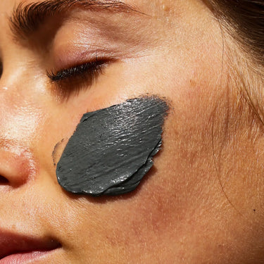Eine Nahaufnahme des Gesichts einer Person mit geschlossenen Augen, die eine dicke Schicht DALUMA CLEANSING AKTIVKOHLE FACE MASK mit marokkanischer Lava Heilerde auf der Wange trägt.