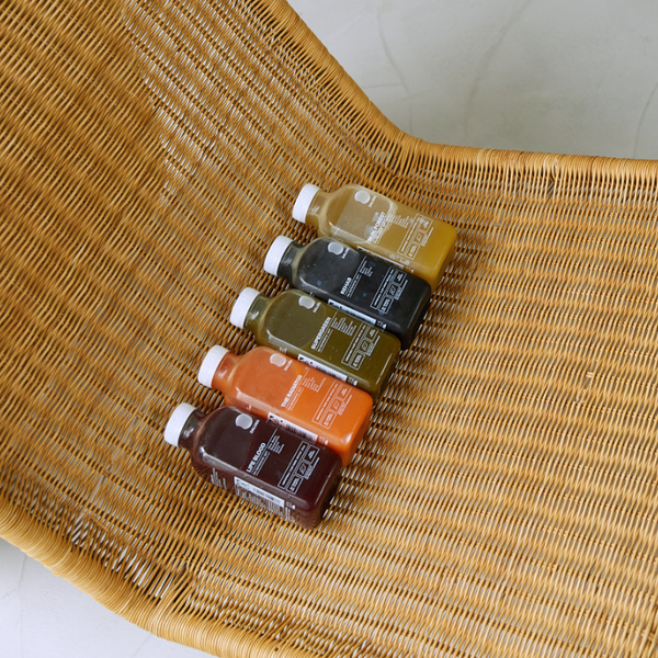 Vier bunte Saftflaschen mit weißen Verschlüssen sind ordentlich in einer Reihe auf einem geflochtenen Rattanstuhl angeordnet. Jede Flasche enthält ein andersfarbiges Getränk, das von dunklen bis hellen Tönen reicht.