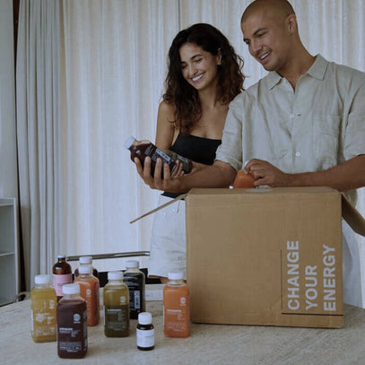 Eine lächelnde Frau und ein lächelnder Mann packen Flaschen mit Säften und Nahrungsergänzungsmitteln aus einem Karton mit der Aufschrift CHANGE YOUR ENERGY auf einem Tisch mit verschiedenen bunten Getränken und Behältern aus.