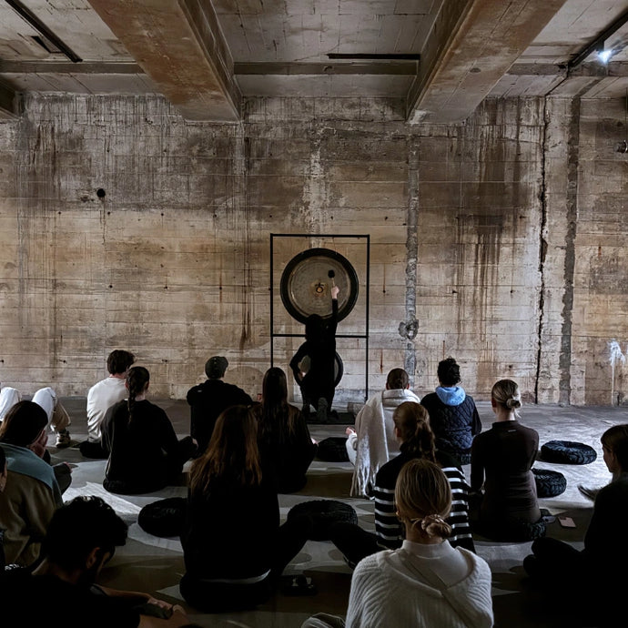 Eine Gruppe von Menschen sitzt im Schneidersitz auf Kissen in einem schummrigen Industrieraum und blickt auf eine Person, die vor einem großen Gong steht, und bereitet sich auf eine Meditations- oder Klangheilungssitzung vor oder nimmt daran teil.