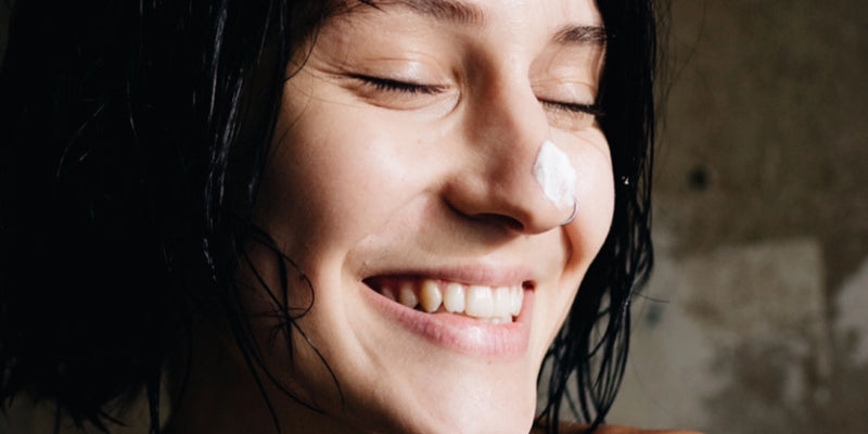 Eine Person mit kurzen dunklen Haaren lächelt mit geschlossenen Augen und zeigt ihre Zähne. An der Nasenspitze befindet sich ein kleiner Klecks weißer Creme. Der Hintergrund ist neutral und unscharf.