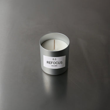 Ein silberner Nullpunkt DUFTKERZE GRADE 0.3 REFOCUS mit einem einzelnen zentrierten Docht ruht unbeleuchtet auf einer glatten grauen Oberfläche - perfekt für geistige Klarheit.