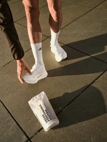 Eine Person, die weiße Adidas-Socken und weiße Turnschuhe trägt, bückt sich, um einen Schuh anzupassen. Ein Paket mit der Aufschrift "Vegan Performance Protein" liegt vor ihr auf dem Bürgersteig. Das Sonnenlicht wirft Schatten auf den Boden.