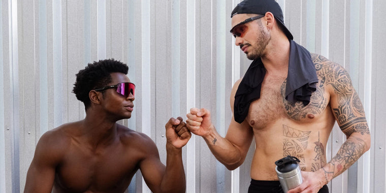 Zwei Männer ohne Hemd und mit Sonnenbrille stoßen sich vor einer Metallwand die Fäuste. Der eine sitzt, der andere steht, hält eine Wasserflasche und hat ein Hemd über der Schulter. Beide wirken sportlich und entspannt.