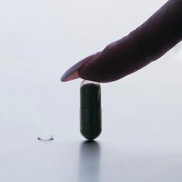 Eine Hand mit polierten Nägeln drückt sanft auf eine DALUMA IMMUN DEFENCE COMPLETE KOMPLEX DOUBLE Kapsel - reich an Kalmeghblattextrakt - die auf einer hellen Fläche neben einem Wassertropfen steht, um das Immunsystem zu stärken.