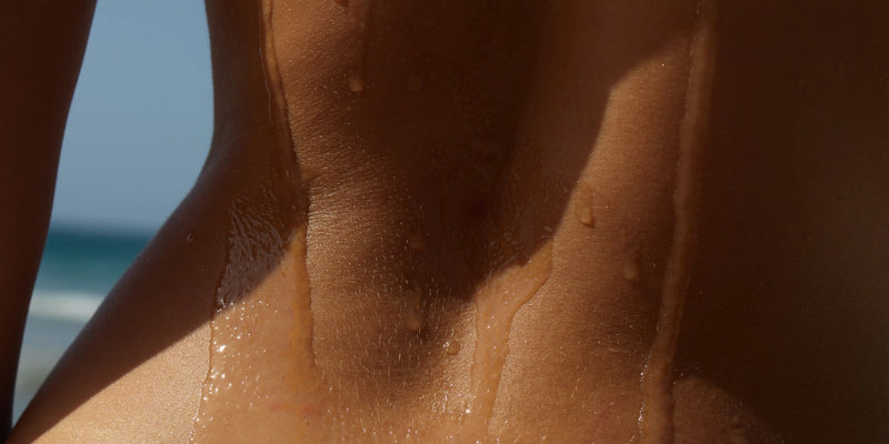 Nahaufnahme einer Person mit glitzernden Wassertropfen auf dem Rücken vor einem unscharfen Hintergrund aus Strand und Meer.