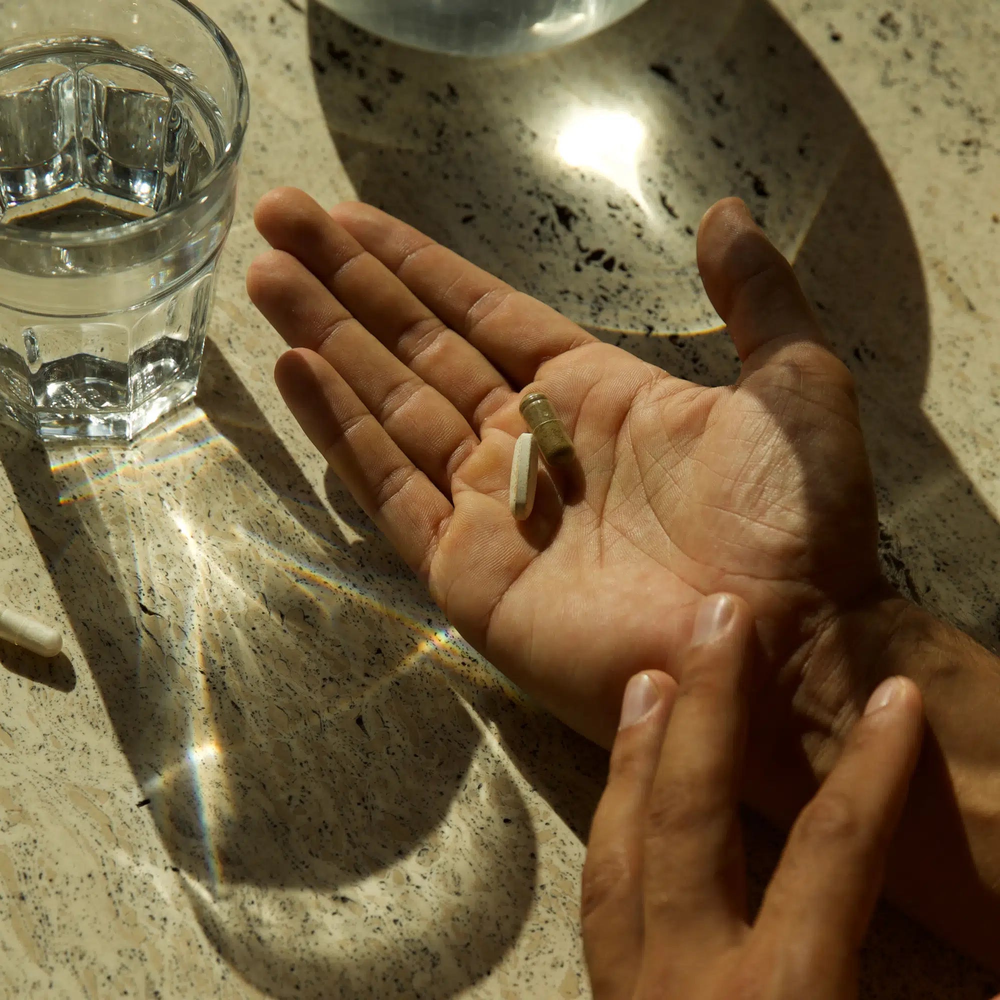 Eine Person hält drei DALUMA DEFENCE DUO Kapseln in ihrer offenen Handfläche neben einem Glas Wasser auf einer sonnenbeschienenen, gesprenkelten Arbeitsplatte. Das Licht des Glases reflektiert auf der Oberfläche und symbolisiert die tägliche Unterstützung des Immunsystems.