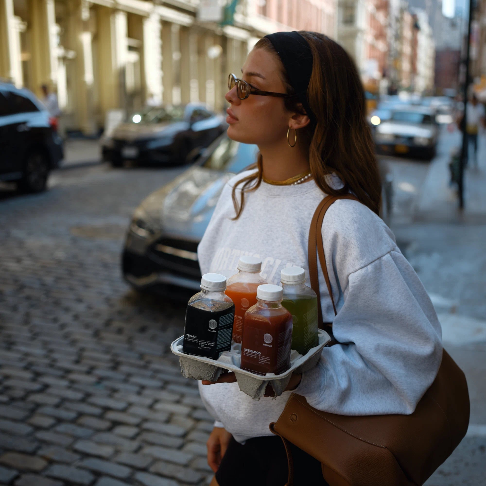 Eine Frau mit Sonnenbrille und Sweatshirt läuft über eine kopfsteingepflasterte Straße in der Stadt und trägt die BESSERE SAFTKUR 7 TAGE Säfte von DALUMA. Mit ihrer braunen Umhängetasche und dem schwarzen Stirnband sieht sie aus, als wäre sie bereit, ihre Reinigung zu beginnen.