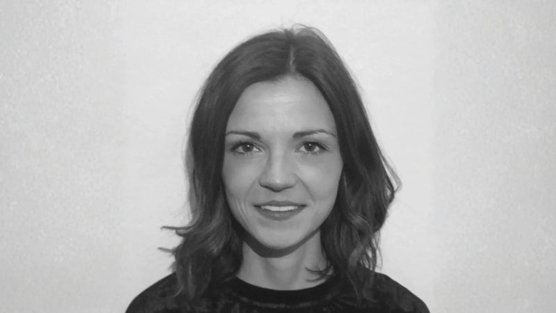 Eine Frau mit schulterlangem, gewelltem Haar, die sanft lächelt und ein dunkles Oberteil trägt, steht vor einem schlichten hellen Hintergrund. Das Foto ist in schwarz-weiß gehalten.