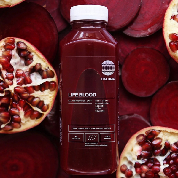Eine Flasche Daluma Life Blood Saft steht zwischen halbierten Granatäpfeln, die seine tiefrote Farbe zur Schau stellen. Auf dem Etikett sind Zutaten wie Rüben, Granatäpfel, Äpfel, Karotten und Cayenne aufgeführt.