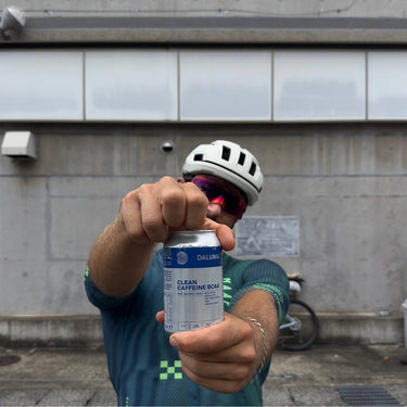 Ein Radfahrer mit weißem Helm und Sonnenbrille hält eine Dose DALUMA CLEAN BCAA ENERGY DRINK 6 PACK hoch, die sein Gesicht teilweise verdeckt. Im Hintergrund ist ein Fahrrad zu sehen, das gegen eine Betonwand gelehnt ist.