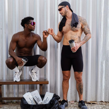 Zwei sportliche Männer in schwarzen Shorts, die Wasserflaschen in der Hand halten, stoßen sich vor einer Metallwand die Fäuste. In der Nähe befinden sich eine Sporttasche und weiße Beutel mit DALUMA VEGANES PERFORMANCE PROTEIN KAKAO, die ihre Wahl eines veganen Proteins unterstreichen.