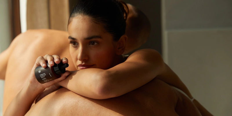 Eine Frau stützt ihr Kinn auf den Armen auf dem Rücken einer anderen Person ab und hält ein dunkles Fläschchen mit einer Pipette. Sie hat ihr dunkles Haar zurückgebunden und blickt in einem schwach beleuchteten Raum nachdenklich zur Seite.