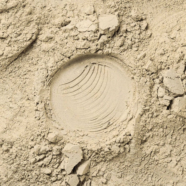 Ein einzelner, teilweise kreisförmiger Abdruck mit Rillenlinien in feinem, hellem Sand, umgeben von losen, ungleichmäßigen Klumpen und verstreuten Körnern.