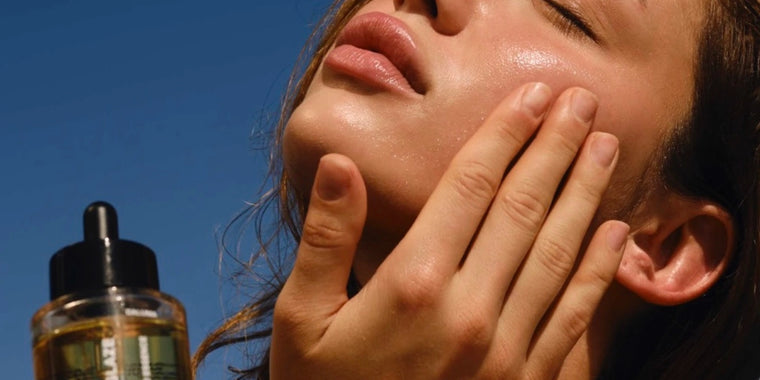 Eine Person trägt mit einer Hand ein Hautpflegeserum auf ihre Wange auf, die Augen geschlossen, unter hellem natürlichem Licht. Eine Tropfflasche mit dem Serum ist seitlich vor einem klaren blauen Himmel zu sehen.