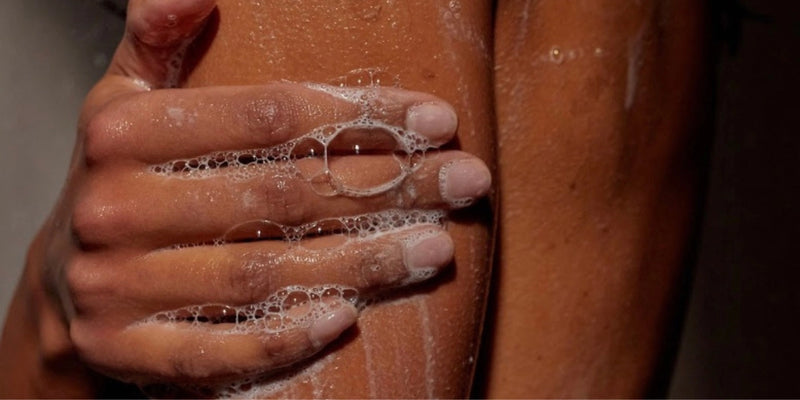 Eine Nahaufnahme einer mit Seifenlauge bedeckten Hand, die sanft einen Arm wäscht, wobei Wassertropfen auf der Haut sichtbar sind. Die Beleuchtung hebt die Textur der Haut und der Seifenblasen hervor.