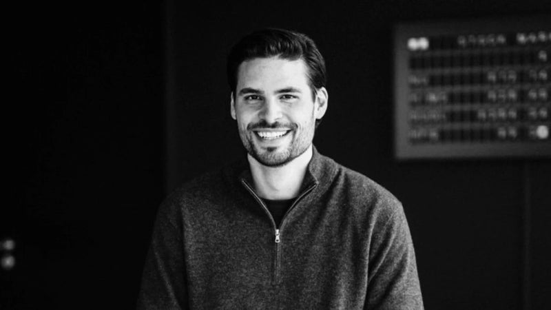 Ein Mann mit kurzen dunklen Haaren und einem Bart sitzt lächelnd in einem Haus. Er trägt einen dunklen Pullover und befindet sich vor einem dunklen Hintergrund mit einem unscharfen Schild an der Wand. Das Bild ist schwarz-weiß.