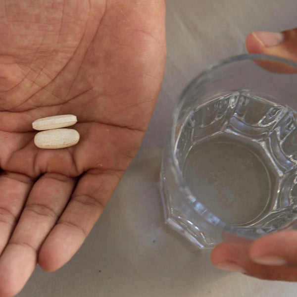 Eine Hand hält zwei ovale weiße Tabletten neben einer anderen Hand, die ein Glas Wasser auf einer hellen Fläche hält.