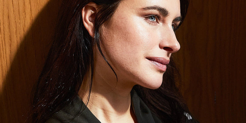 Eine Frau mit langen dunklen Haaren und heller Haut ist in Großaufnahme zu sehen, die nachdenklich zur Seite blickt. Warmes Sonnenlicht beleuchtet ihr Gesicht vor einem hölzernen Hintergrund.