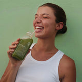 Eine lächelnde Frau in einem weißen Tank-Top nippt mit einem Strohhalm an DALUMAs SUPERGREEN GREEN JUICE 6 PACK und steht vor einer hellgrünen Wand.