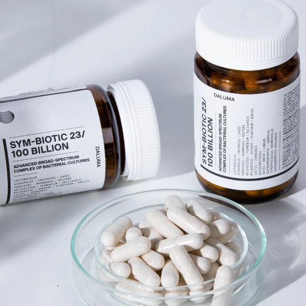 Zwei braune Flaschen mit der Aufschrift SYM-BIOTIC 23/100 BILLION stehen auf einer weißen Fläche, eine aufrecht und eine auf der Seite, neben einer klaren Schale mit weißen Kapseln.