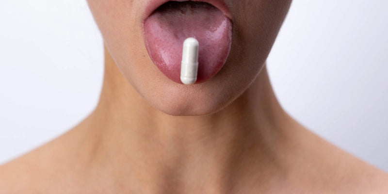 Eine Person mit nackten Schultern streckt ihre Zunge heraus, auf der eine große weiße Kapsel vor einem schlichten weißen Hintergrund ruht.