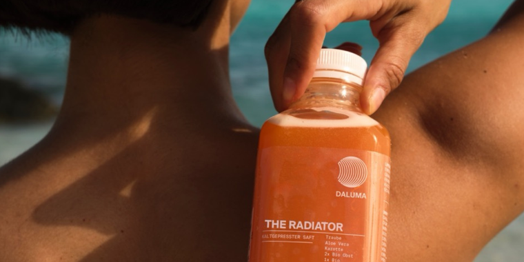 Eine Person hält eine Flasche Daluma The Radiator-Saft dicht an ihre nackte Schulter, während das Meer und der Himmel im Hintergrund verschwimmen.