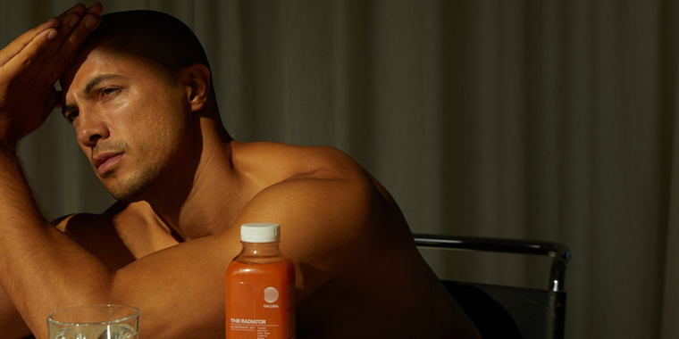 Ein Mann ohne Hemd sitzt an einem Tisch, stützt seinen Kopf auf die Hand und schaut nachdenklich. Vor ihm stehen eine Flasche mit einem orangefarbenen Getränk und ein Glas mit Eiswasser. Der Hintergrund ist durch geschlossene Vorhänge schwach beleuchtet.