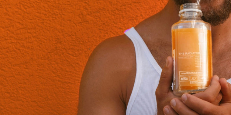 Eine Person in einem weißen Tank-Top hält eine durchsichtige Orangensaftflasche mit der Aufschrift The Radiator neben ihr Gesicht und steht vor einer leuchtend orangefarbenen, strukturierten Wand.