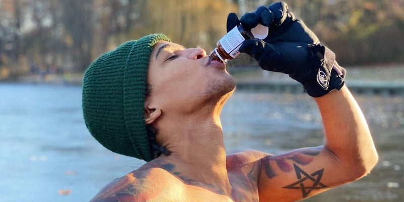 Eine Person ohne Hemd, mit grüner Mütze und schwarzen Handschuhen, trinkt aus einer kleinen Flasche im Freien in der Nähe eines Gewässers, wobei die Bäume im Hintergrund verschwimmen. Auf dem Oberarm ist ein tätowierter Stern zu sehen.