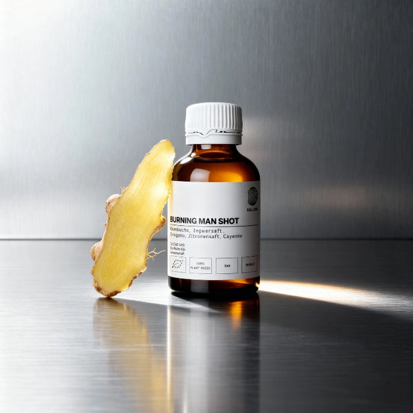 Eine kleine dunkle Flasche mit der Aufschrift Burning Man Shot steht neben einer frischen Scheibe Ingwer auf einer spiegelnden Metalloberfläche mit einem Hintergrund aus gebürstetem Stahl.