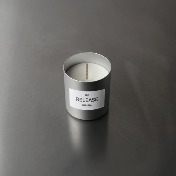 Eine silberne Kerze mit einem weißen Etikett mit der Aufschrift 0.2 RELEASE DALUMA steht auf einer glatten, dunkelgrauen Oberfläche. Die Kerze ist nicht angezündet, und das minimalistische Design sorgt für ein modernes, klares Aussehen.