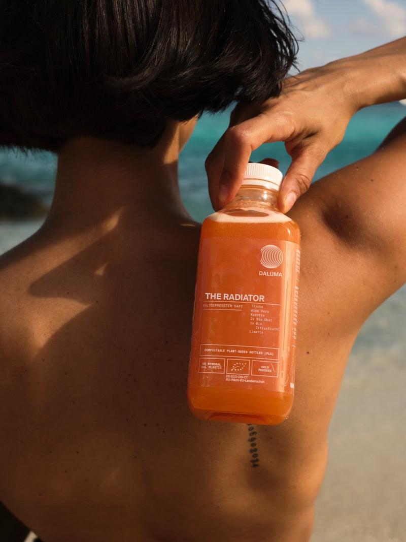 Eine Person mit kurzen dunklen Haaren hält eine Orangensaftflasche mit der Aufschrift THE RADIATOR von DALUMA an ihren nackten Rücken, während sie im Freien an einem Strand mit türkisfarbenem Wasser steht.