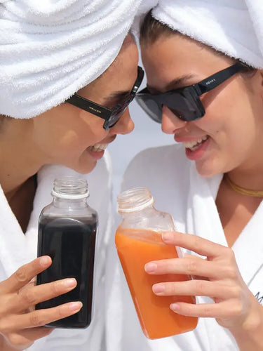 Zwei Frauen in weißen Bademänteln und mit Handtüchern auf dem Kopf lächeln und stoßen mit Sonnenbrillen auf Glasflaschen mit Saft an, eine dunkle und eine orange.