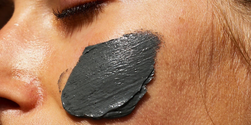 Eine Nahaufnahme der Wange einer Person mit geschlossenen Augen, die eine dicke Schicht einer dunkelgrauen Gesichtsmaske auf der Haut trägt.