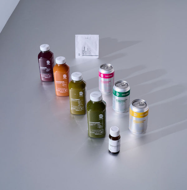 Verschiedene bunte Saftflaschen, Dosen, ein kleines Fläschchen und ein weißes Päckchen sind fein säuberlich auf einer blassgrauen Fläche angeordnet und werfen weiche Schatten nach hinten.