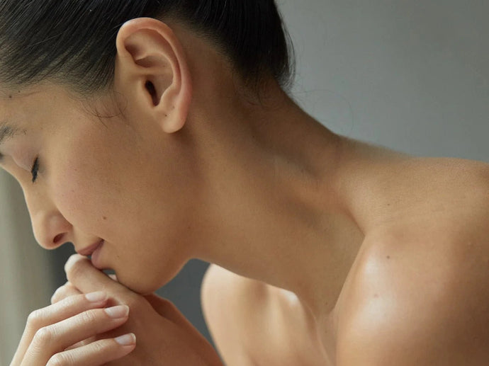 Eine Nahaufnahme einer Frau mit geschlossenen Augen, die ihr Kinn auf die Hände stützt. Sie hat eine glatte Haut, dunkles Haar, das nach hinten gekämmt ist, und wirkt ruhig und entspannt, mit einem neutralen grauen Hintergrund.