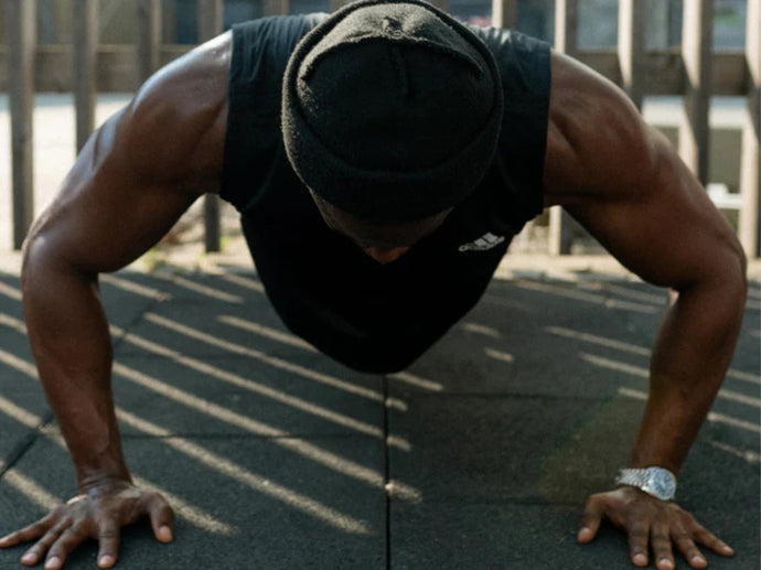 Eine Person, die ein schwarzes ärmelloses Hemd und eine schwarze Mütze trägt, macht Liegestütze im Freien auf einer Gummimatte, wobei das Sonnenlicht Schatten von einem Holzzaun auf ihren Körper wirft.
